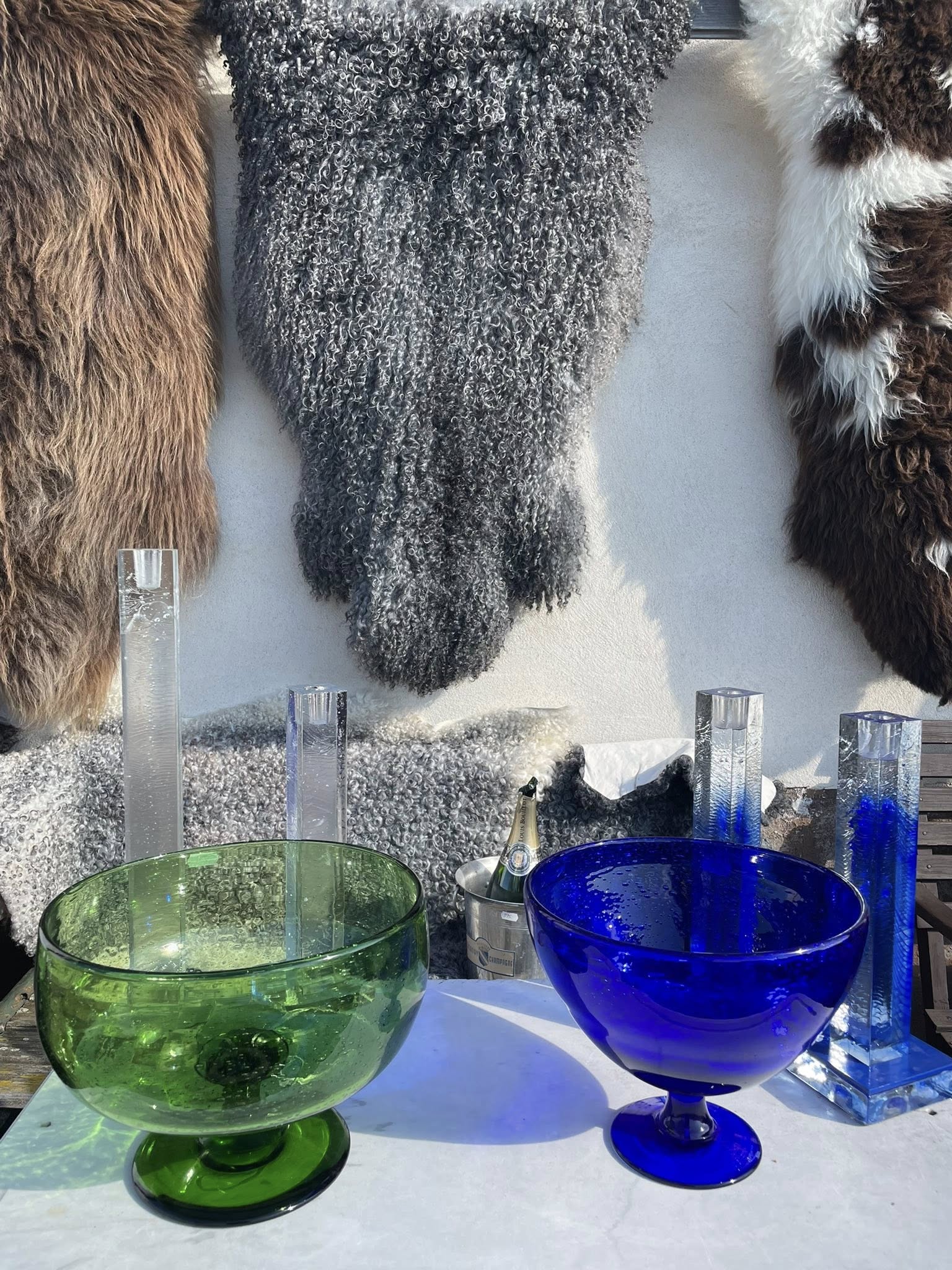 Gröna och blåa glasskålar samt ljusstakar i glas på ett vitt bord med fårskinn i bakgrunden